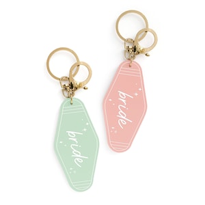 May include: Two keychains with a motel key tag design. The tags are pink and green and say "bride" in white script. The keychains are gold.