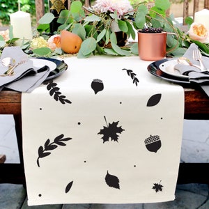 May include: A white table runner featuring a black and white autumn leaf and acorn design. The runner is decorated with leaves, acorns, and small dots.