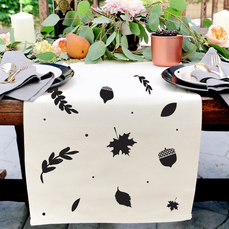 May include: A white table runner featuring a black and white autumn leaf and acorn design. The runner is decorated with leaves, acorns, and small dots.