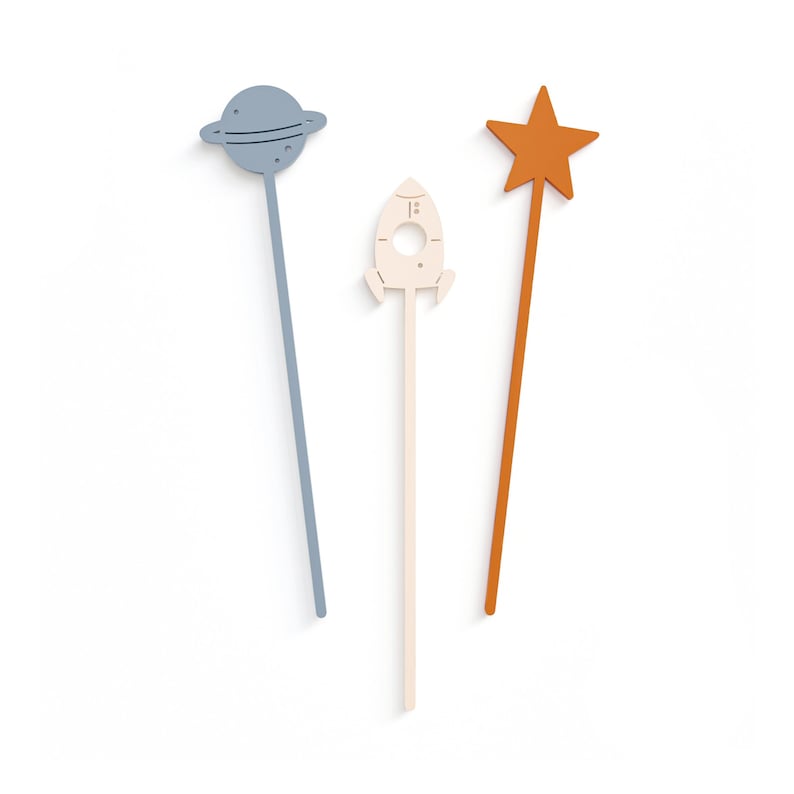 Puede incluir: Tres palitos de madera decorativos con diferentes adornos con tem&aacute;tica espacial: un planeta azul con un anillo, un cohete blanco y una estrella naranja.