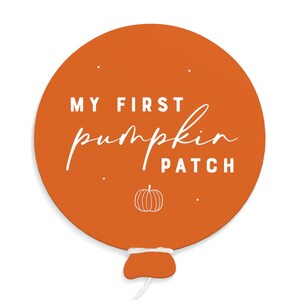 May include: An orange, balloon-shaped sign with the text "MY FIRST pumpkin PATCH" in white. A small pumpkin illustration is at the bottom. The sign has a white string and a small, white and orange accent at the bottom.