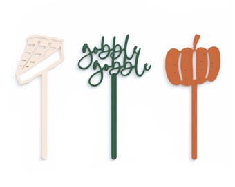 May include: Three wooden Thanksgiving themed cake toppers. The first topper is a slice of pie in beige. The second topper is the words "gobble gobble" in green. The third topper is an orange pumpkin.