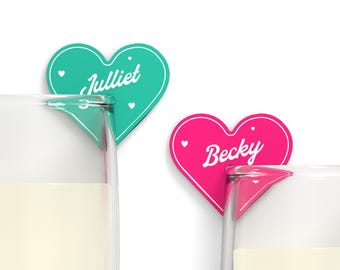 May include: Two heart-shaped glass markers in teal and hot pink. The teal marker features the white text Julliet, and the pink marker features the white text Becky. Both markers have small white heart accents and attach to the rim of a glass.