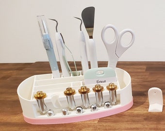 Puede incluir: Organizador de plástico blanco con ribete rosa para guardar herramientas Cricut. Incluye una variedad de herramientas, como una espátula, tijeras, una herramienta de deshierbe y varias cuchillas diferentes. El organizador también tiene un compartimento para guardar un bolígrafo Cricut.
