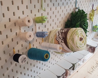 May include: A white pegboard with various spools of thread and yarn in different colors, including white, green, blue, and black. There are also small clear containers on a wooden shelf below the pegboard.