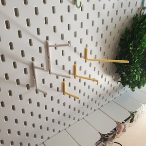 K&ouml;nnte beinhalten: Eine wei&szlig;e Lochwand mit mehreren gelben und grauen Haken. Die Haken sind in verschiedenen Positionen an der Lochwand befestigt. Die Lochwand ist an einer Wand montiert.