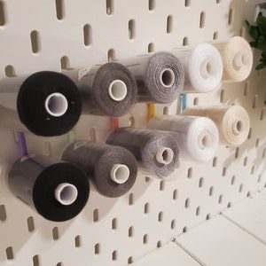 K&ouml;nnte beinhalten: Eine wei&szlig;e Lochwand mit Garnrollen in verschiedenen Farben, darunter Schwarz, Grau, Wei&szlig; und Beige. Die Rollen werden mit Plastikclips befestigt.