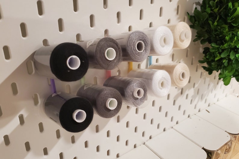 K&ouml;nnte beinhalten: Eine wei&szlig;e Lochwand mit Garnrollen in verschiedenen Farben, darunter Schwarz, Grau, Wei&szlig; und Beige. Die Rollen werden mit Plastikclips befestigt.