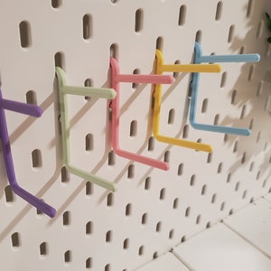 K&ouml;nnte beinhalten: Sechs bunte Kunststoffhaken, die an einer wei&szlig;en Lochwand befestigt sind. Die Haken sind in verschiedenen Pastellfarben gehalten, darunter Lila, Gr&uuml;n, Rosa, Gelb und Blau.