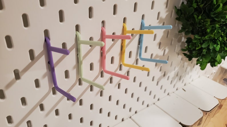 K&ouml;nnte beinhalten: Sechs bunte Kunststoffhaken, die an einer wei&szlig;en Lochwand befestigt sind. Die Haken sind in verschiedenen Pastellfarben gehalten, darunter Lila, Gr&uuml;n, Rosa, Gelb und Blau.