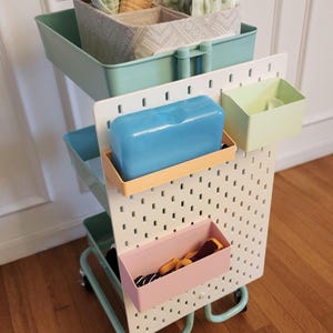 May include: A mint green rolling cart with multiple storage levels. The top level holds fabric items in patterned bins. A white pegboard with attached bins and a blue rectangular container is on the cart.