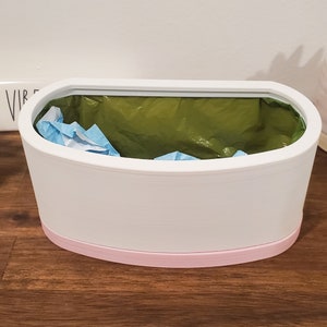 May include: White oval-shaped trash can with a pink rim. The trash can has a green liner and a blue paper towel inside.