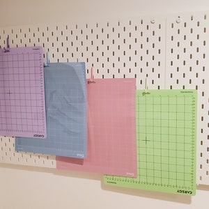 Puede incluir: Cuatro esteras de corte autocicatrizantes en diferentes colores, morado, azul, rosa y verde, están colgadas en una pared blanca de tablero de clavijas. Las esteras son todas del mismo tamaño y tienen un patrón de cuadrícula.