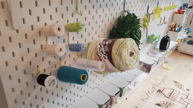 K&ouml;nnte beinhalten: Eine wei&szlig;e Lochwand mit verschiedenen Garnrollen und Wollkn&auml;uel in verschiedenen Farben, darunter wei&szlig;, gr&uuml;n, blau und schwarz. Unter der Lochwand befindet sich ein Holzregal mit kleinen durchsichtigen Beh&auml;ltern.