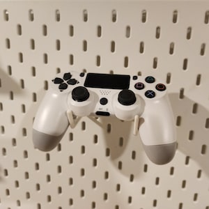 Pode incluir: Um controlador de videogame sem fio branco com botões pretos e uma tela preta. O controlador está pendurado em uma placa de pinos branca.