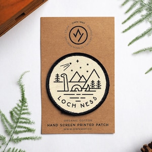 Könnte beinhalten: Ein runder, weißer, handbedruckter Patch mit schwarzem Rand. Der Patch zeigt eine schwarze Linienzeichnung eines Seeungeheuers im Loch Ness in einem See mit Bergen und Sternen im Hintergrund. Der Text "Loch Ness" ist in Schwarz unter dem Bild gedruckt. Der Patch befindet sich auf einer braunen Karte mit dem in Schwarz gedruckten Text "Own Way Graphic Design Screen Printing".