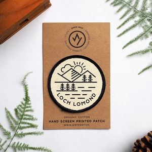 Peut inclure: Un patch brodé blanc et noir avec une chaîne de montagnes, un soleil et des arbres. Le texte "Loch Lomond" est imprimé sur le patch. Le patch est sur une carte brune avec le texte "Organic Cotton Hand Screen Printed Patch www.ownway.co".