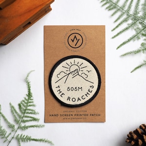 Puede incluir: Un parche redondo blanco impreso a mano con contornos negros. El parche presenta una montaña con un sol, un pájaro y el texto "The Roaches 505M".