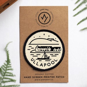 Könnte beinhalten: Ein runder Aufnäher mit schwarzem Rand und cremefarbenem Zentrum. Er zeigt eine schwarze Illustration einer Küstenstadt und eines Bootes, mit dem Wort "ULLAPOOL" in Schwarz gedruckt. Der Aufnäher befindet sich auf einer braunen Karte mit Text und Logo.