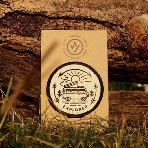 May include: A beige embroidered patch with a black outline depicting a vintage camper van with a pop-up top, a sun, and trees. The text "EXPLORER" is below the camper van. The patch is on a brown paper background.