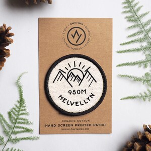 May include: A white, round, hand-screen printed patch with a black outline. The patch features a mountain range with a sun rising behind it. The text "950M HELVELLYN" is printed in black below the mountains. The patch is made of organic cotton.