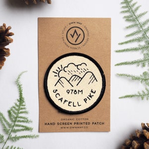 Op de afbeelding: Een handbedrukte patch van witte en zwarte stof met een berglandschap en de tekst "978M Scafell Pike". De patch is gemaakt van biologisch katoen en rond.