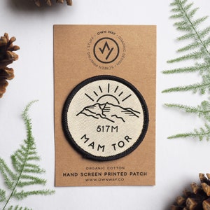 May include: A white and black round patch with a mountain range and sun graphic. The text "517M MAM TOR" is printed on the patch. The patch is made of organic cotton and is hand screen printed.