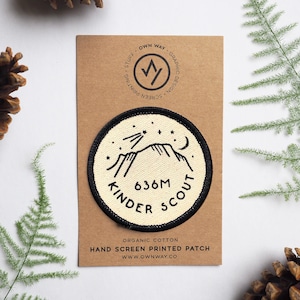 May include: A white, round, embroidered patch with black stitching. The patch features a mountain range with a crescent moon and stars. The text "636M KINDER SCOUT" is written in a black circle around the mountain range. The patch is on a brown card with the text "OWN WAY GRAPHIC DESIGN SCREEN PRINTING STUFF" printed on it.