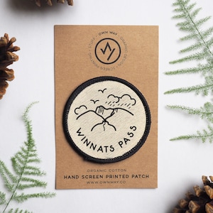 May include: A white, round, hand-screen printed patch with black outlines of a mountain range, birds, and rain clouds. The text "WINNATS PASS" is written in a black, circular font around the image. The patch is on a brown card with the text "OWN WAY GRAPHIC DESIGN SCREEN PRINTING STUFF" in black.