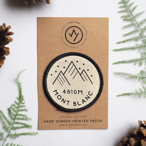 Könnte beinhalten: Ein runder, weißer Stoff-Patch mit schwarzen Nähten und einem schwarzen Bergketten-Design. Der Text "4810M MONT BLANC" ist in Schwarz auf dem Patch gedruckt. Der Patch befindet sich auf einer braunen Karte mit dem in Schwarz gedruckten Text "OWN WAY GRAPHIC DESIGN SCREEN PRINTING STUFF".