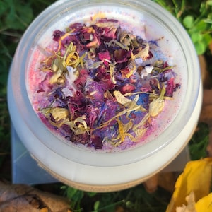 Puede incluir: Una vela blanca con flores secas y purpurina incrustadas en la cera. La vela está en un tarro de vidrio transparente.