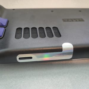 以下が含まれることがあります： 紫色のボタンが付いた黒い携帯型ゲーム機のクローズアップ。6つの長方形の通気孔とホログラフィックステッカーが特徴です。「VALVE」の文字が見えます。