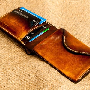 Op de afbeelding: Open, handgemaakte leren portemonnee in bruintinten, met zichtbare stiksels en een gebogen flap. Verschillende creditcards zijn gedeeltelijk zichtbaar in de portemonnee. De portemonnee ligt op een getextureerd, bruin oppervlak.