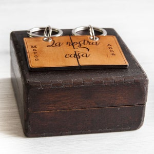 May include: A dark brown wooden box with a light brown, engraved tag that reads "La nostra casa". The tag is split into two pieces and attached to two key rings. The names "Marco" and "Anna" are also engraved on the tag.