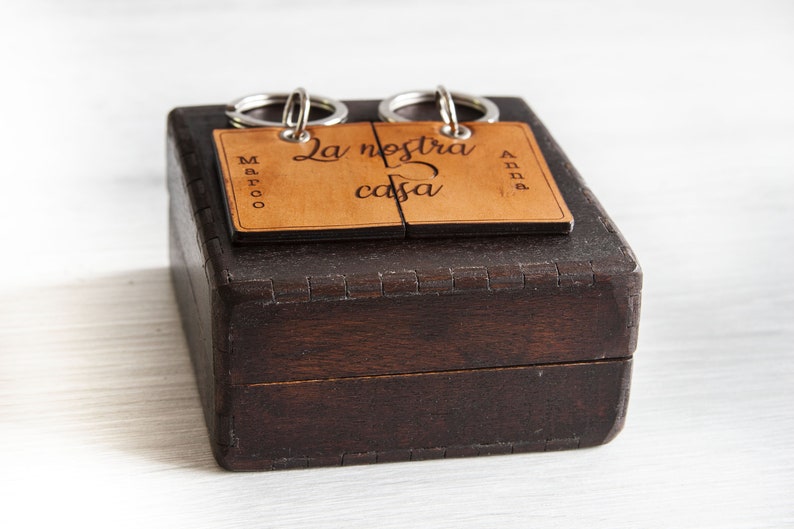 May include: A dark brown wooden box with a light brown, engraved tag that reads "La nostra casa". The tag is split into two pieces and attached to two key rings. The names "Marco" and "Anna" are also engraved on the tag.