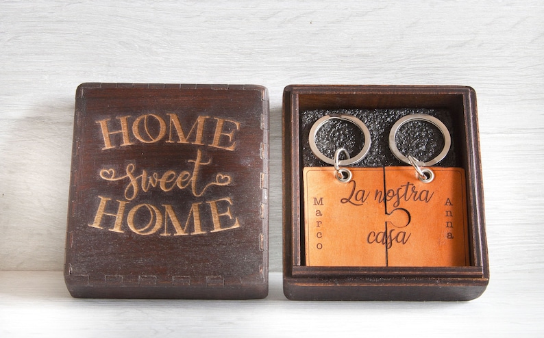 May include: A dark brown wooden box with the words "HOME sweet HOME" engraved on the lid. Inside, two keychains with a puzzle piece design are visible, each with a name and the phrase "La nostra casa".