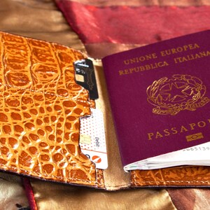 Puede incluir: Un portapasaporte de cuero marr&oacute;n con un patr&oacute;n de cocodrilo. El portapasaporte tiene una ranura para un pasaporte, tarjetas de cr&eacute;dito y otros documentos. El portapasaporte est&aacute; abierto y un pasaporte italiano burdeos es visible en el interior.