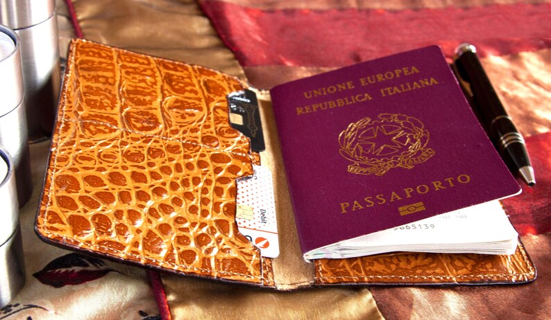 Puede incluir: Un portapasaporte de cuero marr&oacute;n con un patr&oacute;n de cocodrilo. El portapasaporte tiene una ranura para un pasaporte, tarjetas de cr&eacute;dito y otros documentos. El portapasaporte est&aacute; abierto y un pasaporte italiano burdeos es visible en el interior.