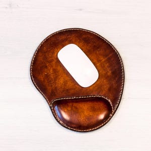 May include: A brown leather mouse pad with a built-in wrist rest and a white wireless mouse. The mouse pad has a stitched border and a smooth, polished surface. The wrist rest is a darker shade of brown.