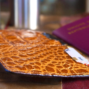 Puede incluir: Un portapasaporte de cuero marr&oacute;n con un patr&oacute;n de cocodrilo. El portapasaporte est&aacute; abierto y un pasaporte es visible en el interior.
