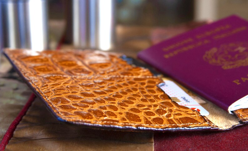 Puede incluir: Un portapasaporte de cuero marr&oacute;n con un patr&oacute;n de cocodrilo. El portapasaporte est&aacute; abierto y un pasaporte es visible en el interior.