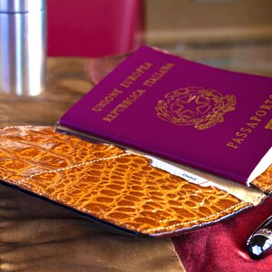 Puede incluir: Un portapasaporte de cuero burdeos con un pasaporte italiano en relieve dorado en el interior. El portapasaporte tiene un patr&oacute;n de cocodrilo y est&aacute; sobre una superficie de tela roja y dorada. Tambi&eacute;n hay un bol&iacute;grafo plateado sobre la superficie.
