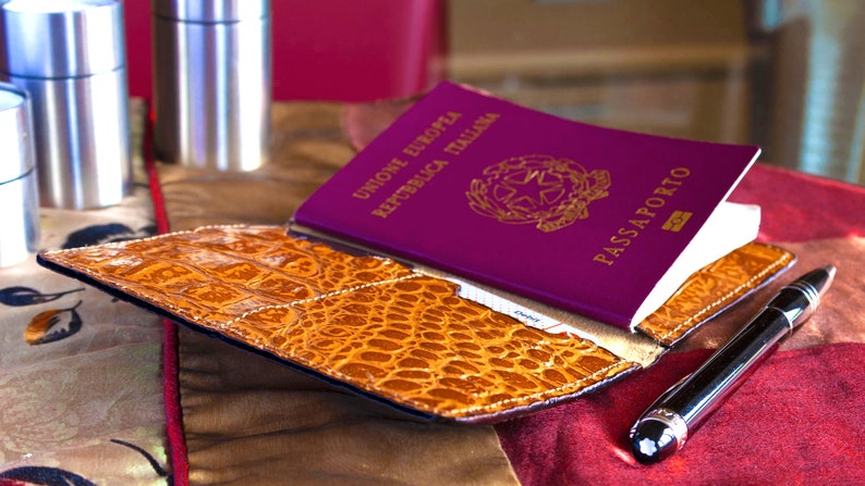Puede incluir: Un portapasaporte de cuero burdeos con un pasaporte italiano en relieve dorado en el interior. El portapasaporte tiene un patr&oacute;n de cocodrilo y est&aacute; sobre una superficie de tela roja y dorada. Tambi&eacute;n hay un bol&iacute;grafo plateado sobre la superficie.