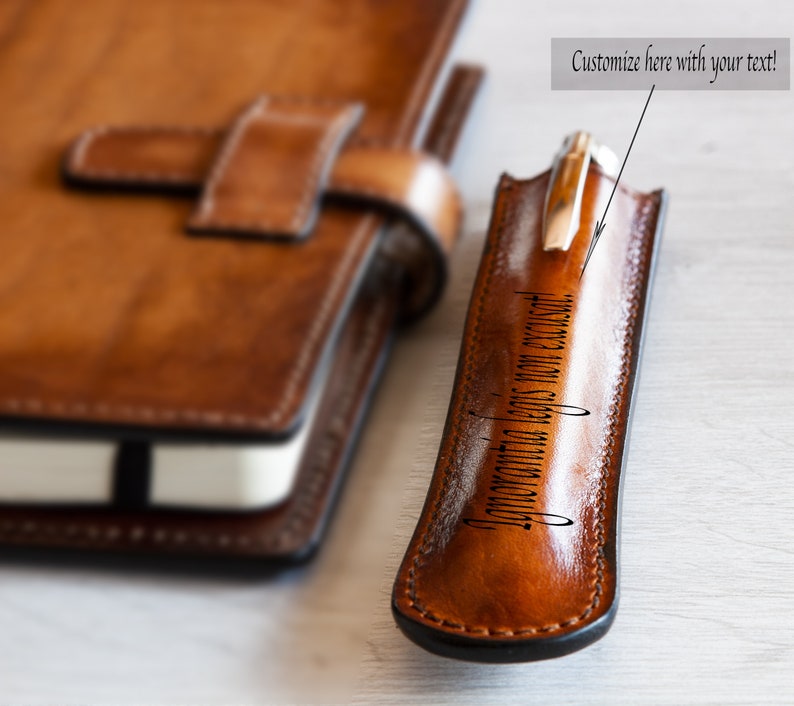 May include: A brown leather pen holder with a silver pen inside. The holder is personalized with the text "Adventure begins on an excuse!".