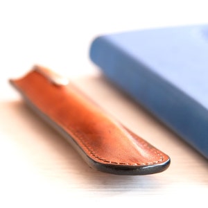 May include: A brown leather pen case with a black stitched edge and a blue notebook in the background.