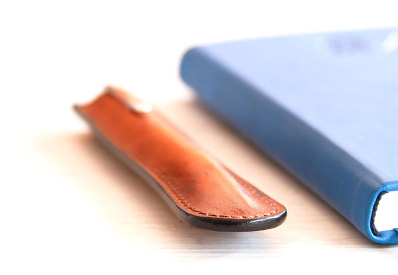 May include: A brown leather pen case with a black stitched edge and a blue notebook in the background.