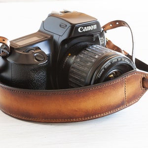 Peut inclure: Un appareil photo Canon EOS Rebel noir avec une sangle en cuir marron. L'appareil photo a un objectif fixé et la sangle est enroulée autour de l'appareil photo.