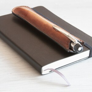 May include: A brown leather pen holder with a silver pen inside, resting on top of a black notebook with a grey ribbon bookmark.