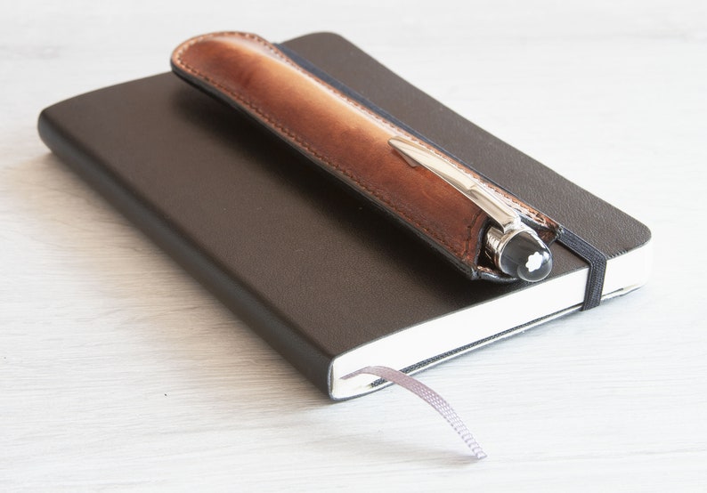 May include: A brown leather pen holder with a silver pen inside, resting on top of a black notebook with a grey ribbon bookmark.