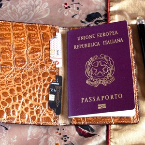 Puede incluir: Un portapasaporte de cuero marr&oacute;n con un pasaporte italiano morado en su interior. El pasaporte tiene el texto "Unione Europea Repubblica Italiana" y "Passaporto" en la portada. Un bol&iacute;grafo plateado est&aacute; sobre una superficie de tela beige junto al portapasaporte.